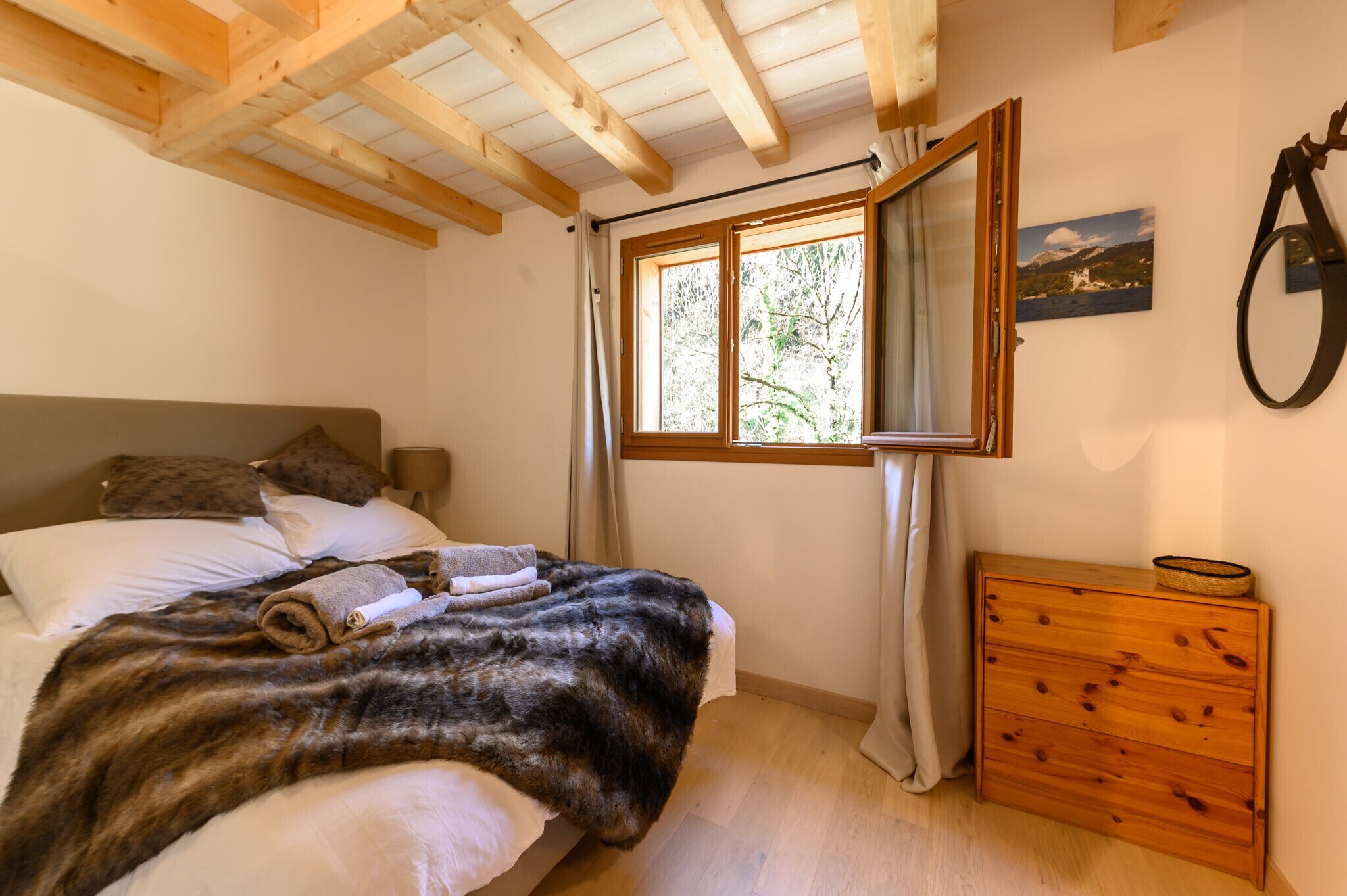Une chambre à coucher avec une tête de lit en bois, une grande fenêtre avec des rideaux, une commode en bois et un tableau accroché au mur.