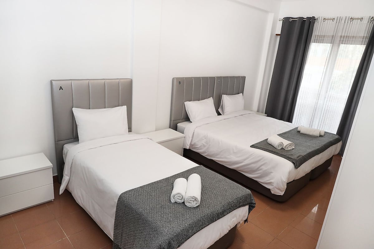 Una habitación de hotel con dos camas individuales, una mesita de noche pequeña y un ventanal grande con cortinas.