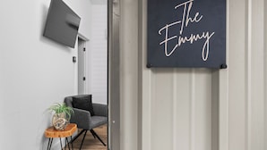 Un couloir avec une enseigne sombre sur le mur indiquant « The Emmy », un fauteuil gris, une petite table en bois avec une plante et une porte moderne.