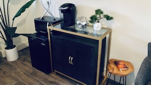 Un espace de vie compact comprenant un petit meuble noir, une machine à café, une plante en pot, un objet décoratif hexagonal et un petit tabouret en bois sur lequel est posé un livre.