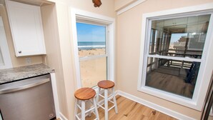 A kitchen with a dishwasher, two stools, a window with a beach view, and a wooden deck outside. - OCEANFRONT, Ocean Views, Screened porch, KH17, Down by the Sea (Kitty Hawk)