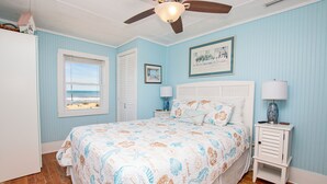 A bedroom with a bed, a ceiling fan, a nightstand, a lamp, a window with a view, and a framed picture on the wall. - OCEANFRONT, Ocean Views, Screened porch, KH17, Down by the Sea (Kitty Hawk)