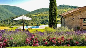 A stone building with a sloped roof, surrounded by a garden with lavender and red flowers, and a view of green hills in the background. - VILLA AMANTA - Nine Bedroom Villa, Sleeps 16 (Perugia)