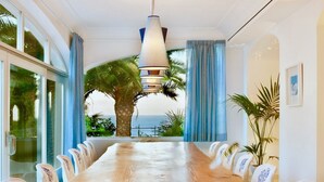 Une longue table de conférence en bois, entourée de chaises blanches, un luminaire suspendu et une vue sur les palmiers et la mer, visible à travers de grandes fenêtres.