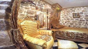 A luxurious room with a stone wall, a tufted gold armchair, a matching ottoman, and a bed with ornate bedding.