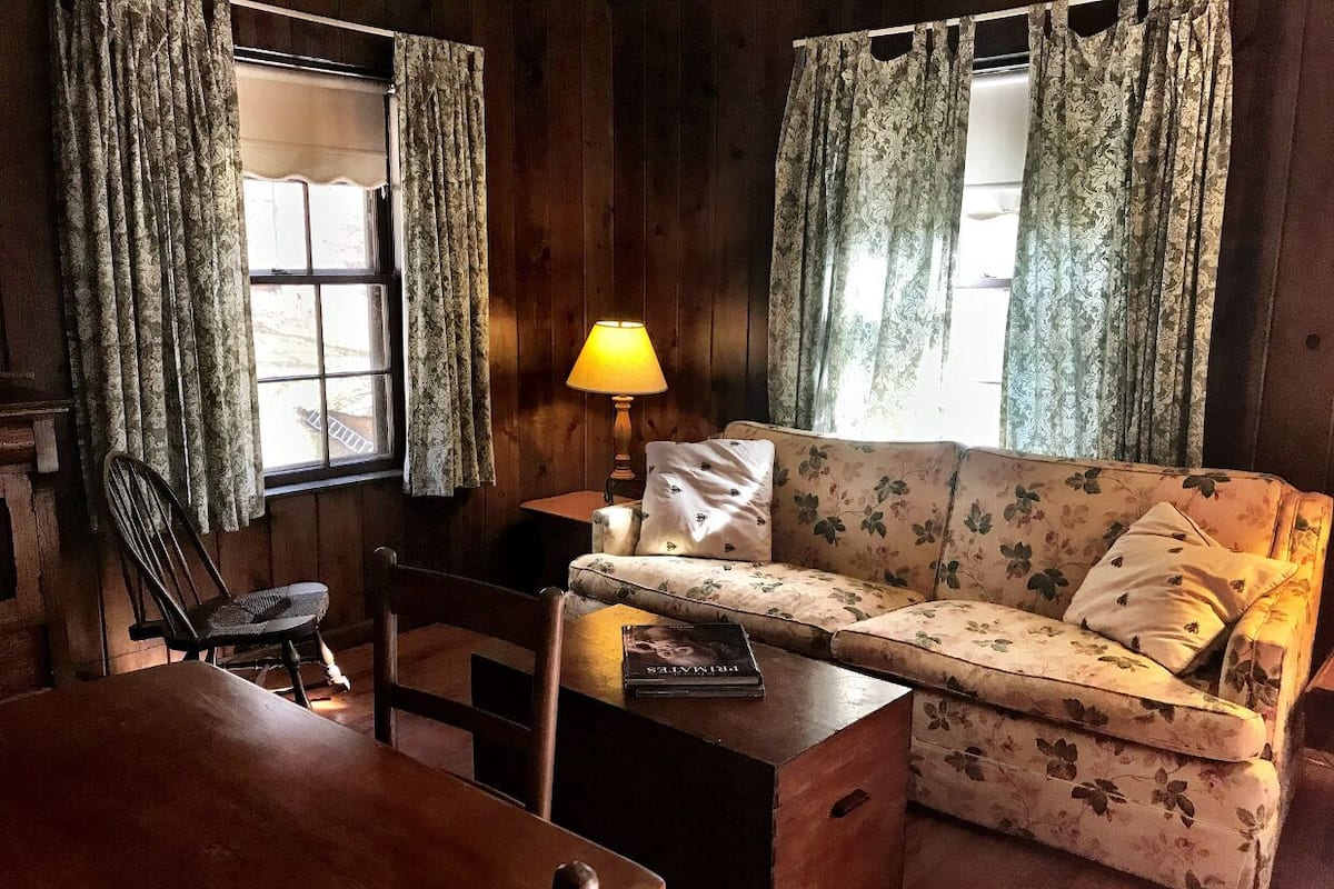 A cozy room with a wooden table, a floral sofa, a chair, and a lamp.