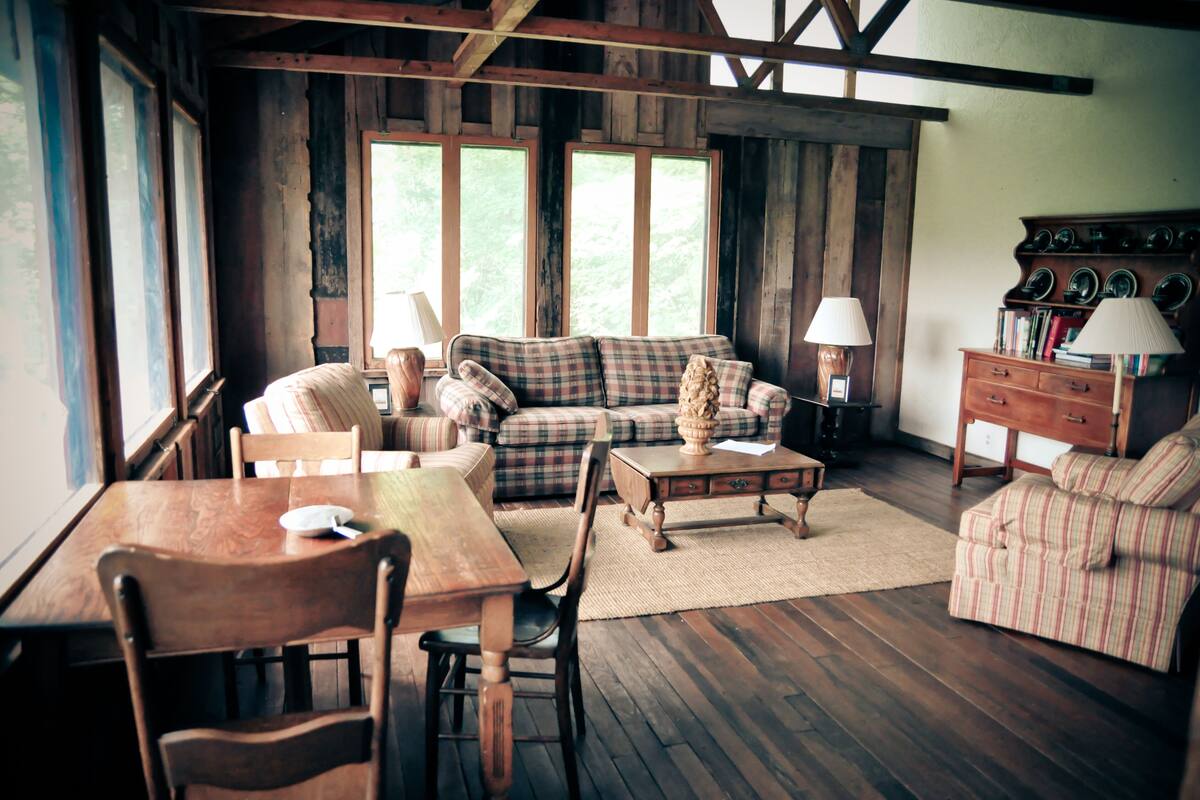 A rustic living room with wooden floors, a plaid sofa, a wooden coffee table, and a dining table with chairs.