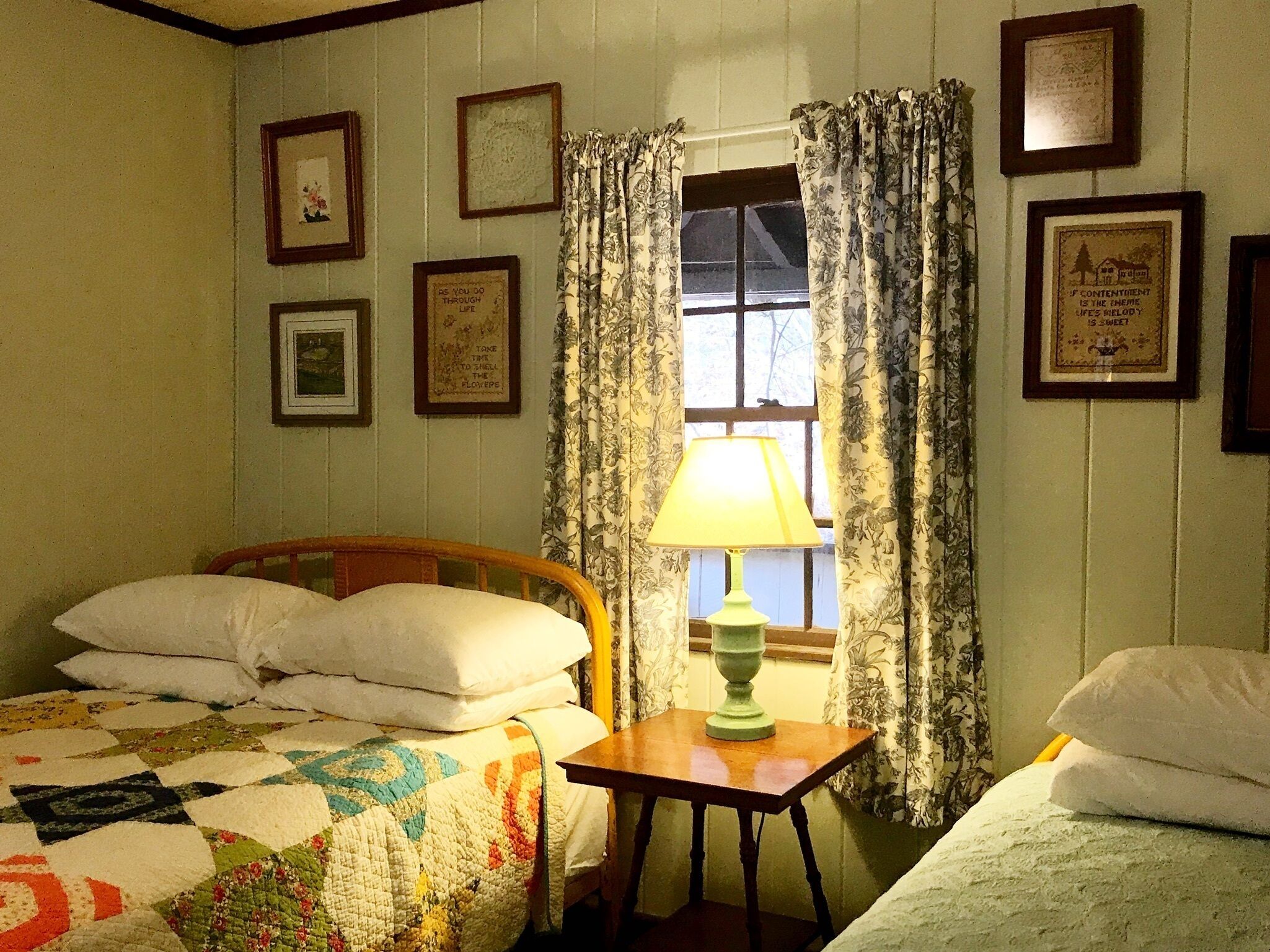 Una habitación con una cama, una mesa con lámpara y una ventana con cortinas. Hay cuadros enmarcados en las paredes.