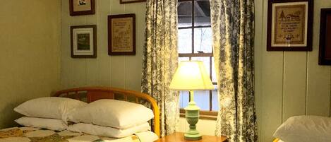 Una habitación con una cama, una mesa con lámpara y una ventana con cortinas. Hay cuadros enmarcados en las paredes.