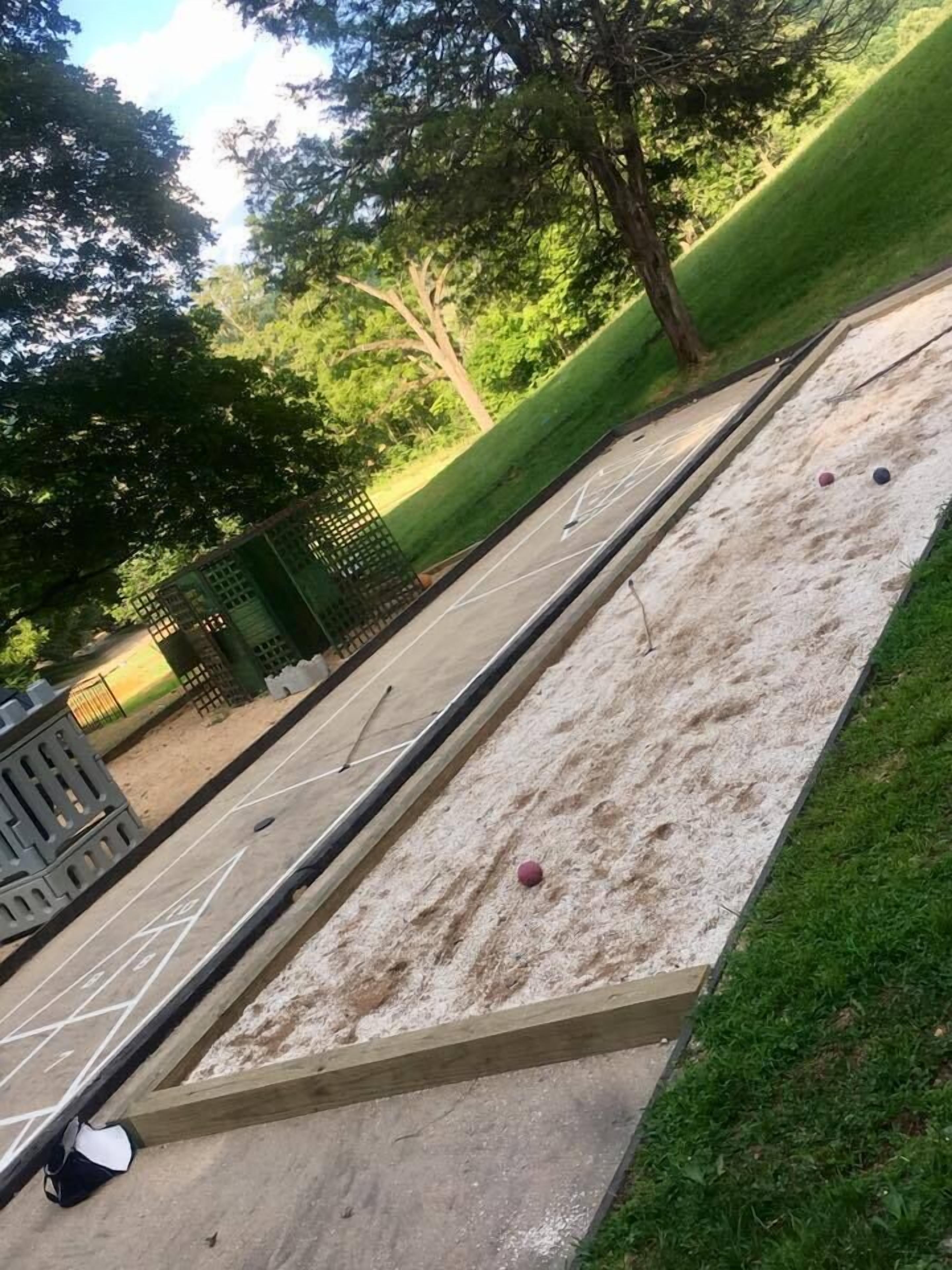 Ein Shuffleboardfeld mit Holzrahmen, einem grünen Zaun und Bäumen im Hintergrund.