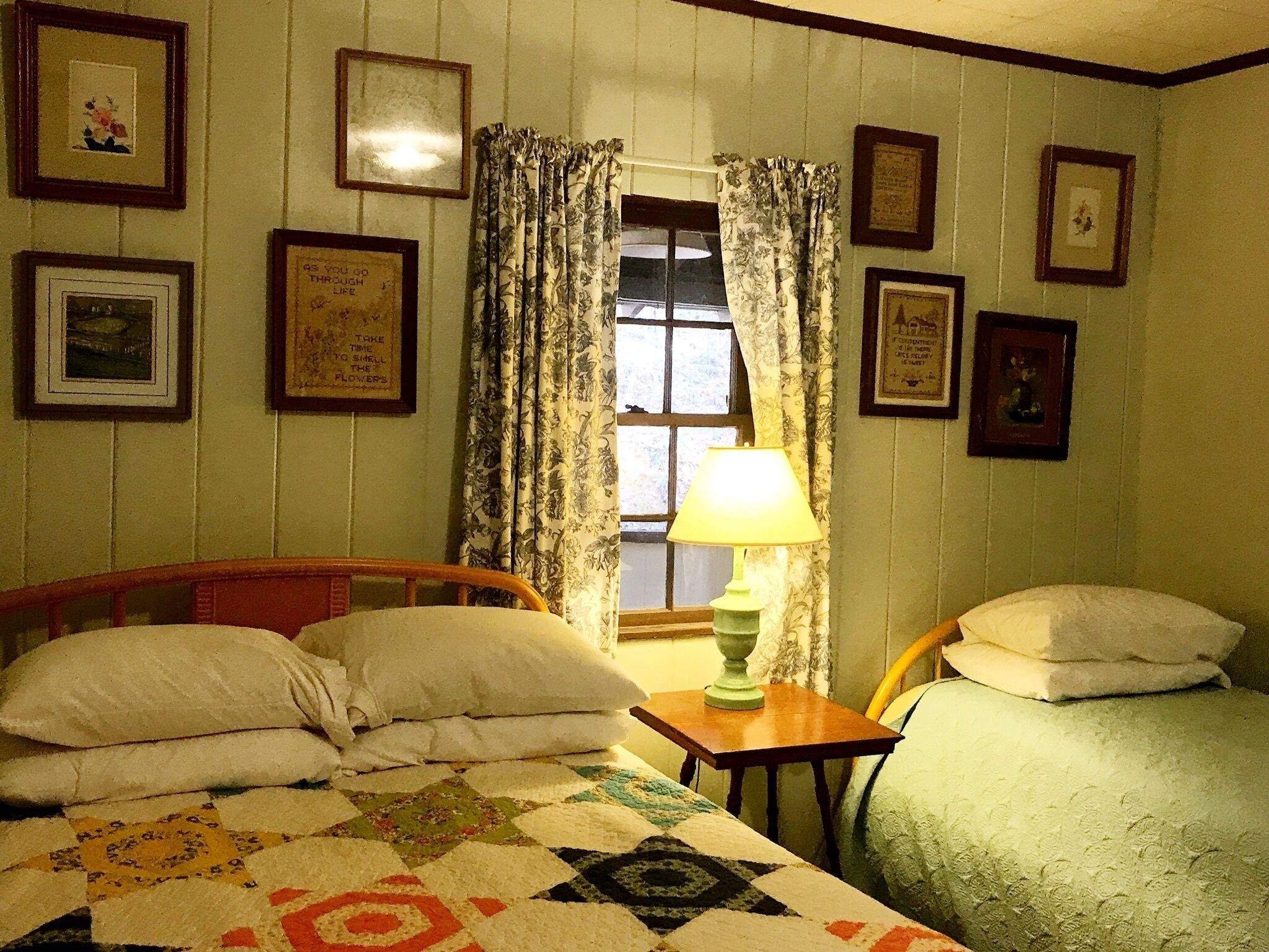 Una habitación acogedora con una cama, una silla y una mesa. Hay cortinas en la ventana y varias fotografías enmarcadas en las paredes.