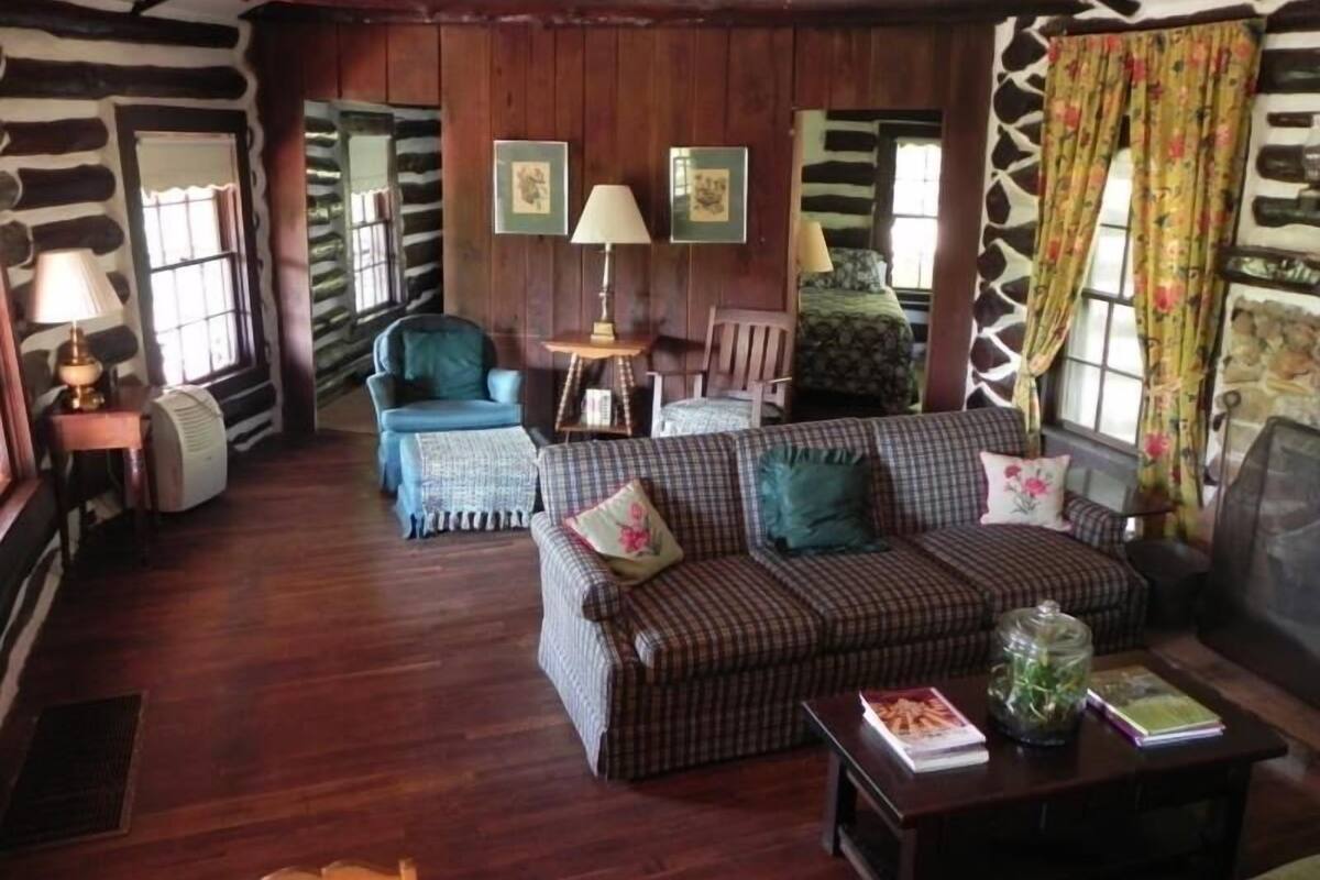 A cozy cabin-style room with a wooden floor, a plaid sofa, a blue armchair, and a small table with a lamp and books.