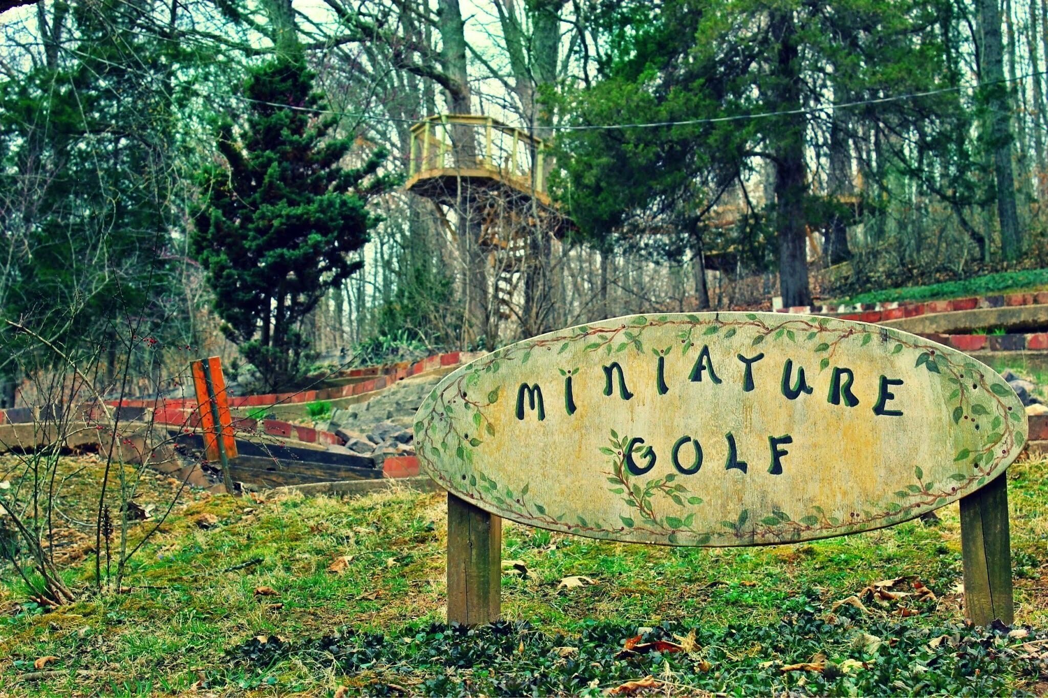 A sign for a miniature golf course surrounded by trees and a wooden structure in the background.