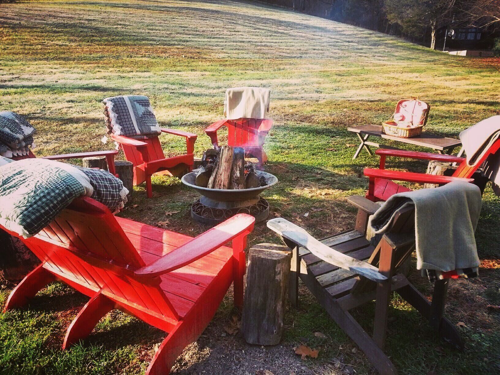 Ein Feuerkorb, umgeben von roten Adirondack-Stühlen mit Decken, in einer grünen, offenen Umgebung.