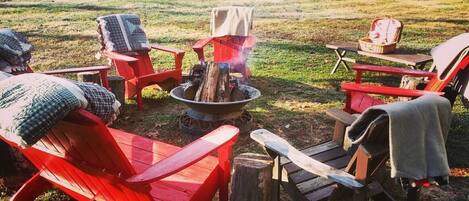 Ein Feuerkorb, umgeben von roten Adirondack-Stühlen mit Decken, in einer grünen, offenen Umgebung.