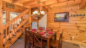 A wooden cabin interior with a dining table set for four, a staircase, and a chandelier. - Smokey Max Cabin sleeps 8, hot tub, jacuzzi , pool table (Pigeon Forge)
