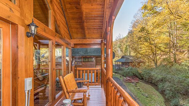 A wooden cabin with a porch, wooden furniture, and a view of the surrounding forest. - Smokey Max Cabin sleeps 8, hot tub, jacuzzi , pool table (Pigeon Forge)