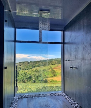 A modern bathroom with a large window, a showerhead, and a view of a green landscape. - Inspire Costa Rica (Alajuela)