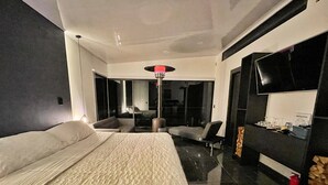 Una habitación de hotel moderna con una cama grande, un televisor de pantalla plana, una zona de estar con sofá y sillones, y una mesa de comedor con un cartel de “Bienvenido”.