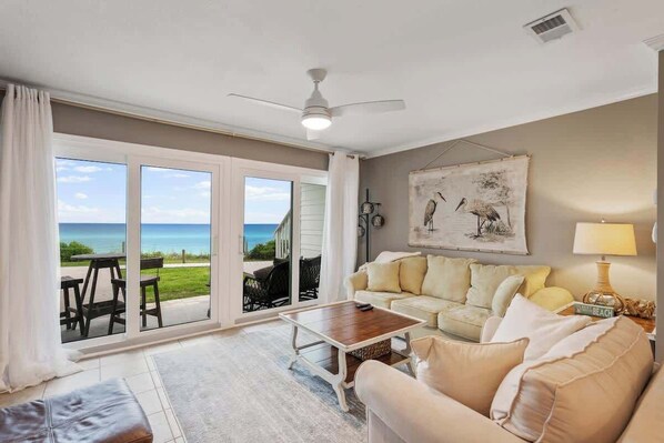 A living room with a large sliding glass door leading to a balcony with a view of the ocean. There is a beige sofa, a wooden coffee table, and a ceiling fan. - Gulf-Front Condo w/Pool & Beach Chair Access (Panama City Beach)