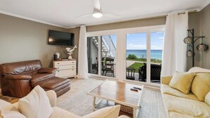 A living room with a brown leather sofa, a white sofa, a coffee table, and a flat-screen TV. There is a sliding glass door leading to a balcony with a view of the ocean. - Blue Tide (Panama City Beach)