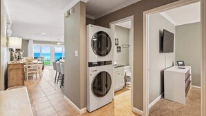 A laundry room with two stacked washers and dryers, a dining area with a view, and a small TV on a white cabinet. - Blue Tide (Panama City Beach)