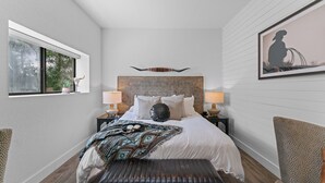 A bedroom with a large bed, two bedside tables with lamps, a bench, and a window with a view of trees. - The Hayes (Bulverde)
