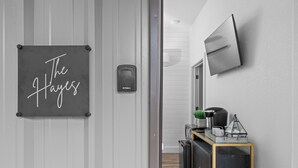 A hallway with a sign reading "The Hayes," a mounted television, and a small kitchenette area with a coffee machine and decorative items. - The Hayes (Bulverde)
