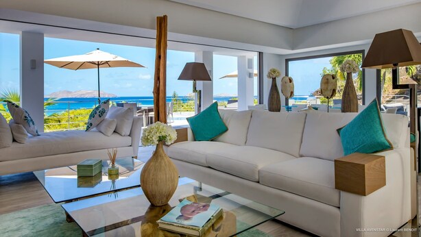 Un salon moderne avec un canapé blanc, une table basse en verre et des objets décoratifs. De grandes fenêtres offrent une vue sur l’océan et les montagnes.