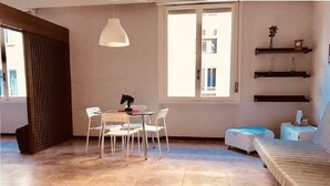Une pièce comprenant un lit, une table à manger avec des chaises, un luminaire suspendu et une fenêtre donnant sur des immeubles.