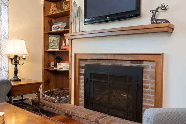 A living room with a fireplace, a television mounted above it, a bookshelf, and a lamp. - Townhome with Fireplace & Hill Views, Saltbox (Hartford)