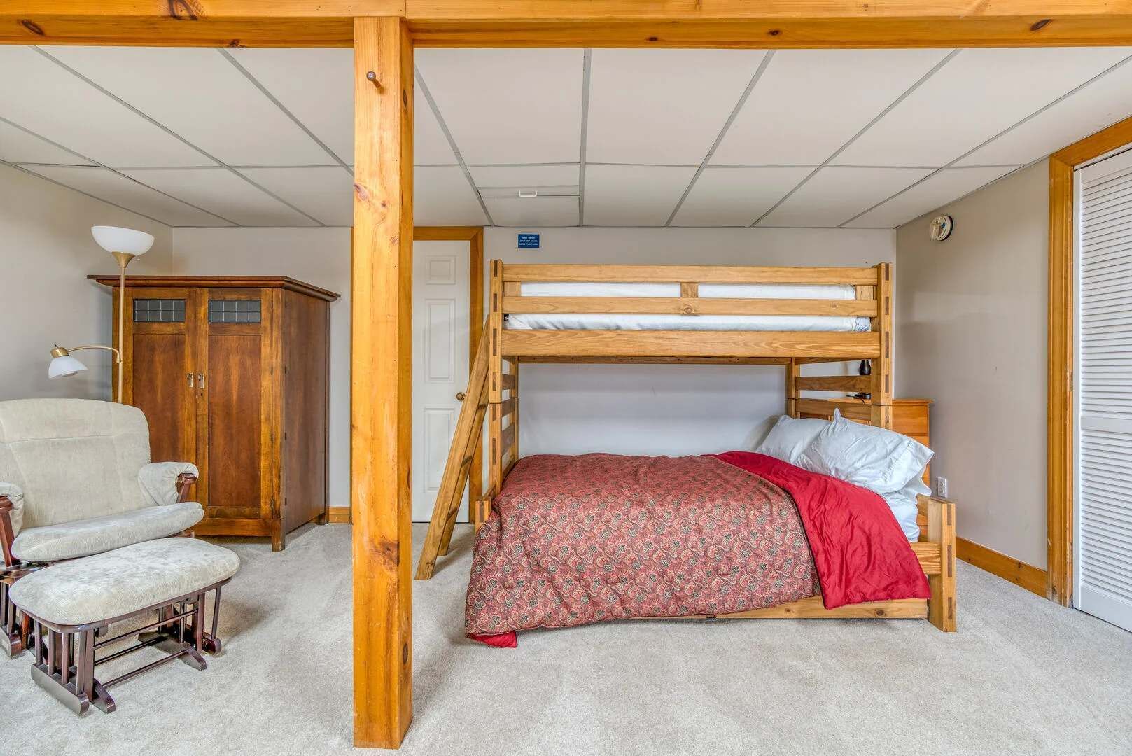 Un lit superposé avec une structure en bois, une chaise, une armoire et une lampe dans une pièce dont le plafond est recouvert de carreaux.