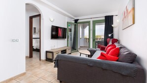 Un saló modern amb un sofà gris, coixins vermells, una televisió plana, una taula baixa de fusta i una porta que dona a un balcó.