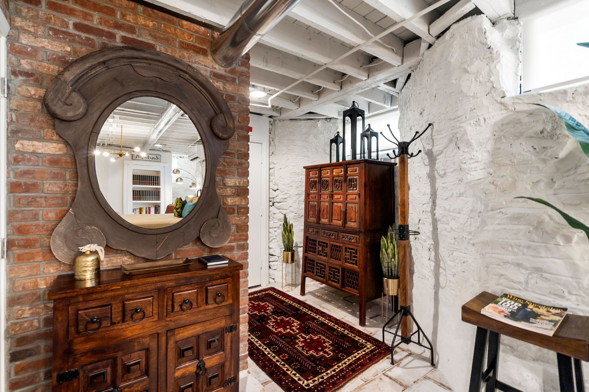 A room with exposed brick walls, a wooden cabinet with drawers, a large mirror, and a patterned rug.