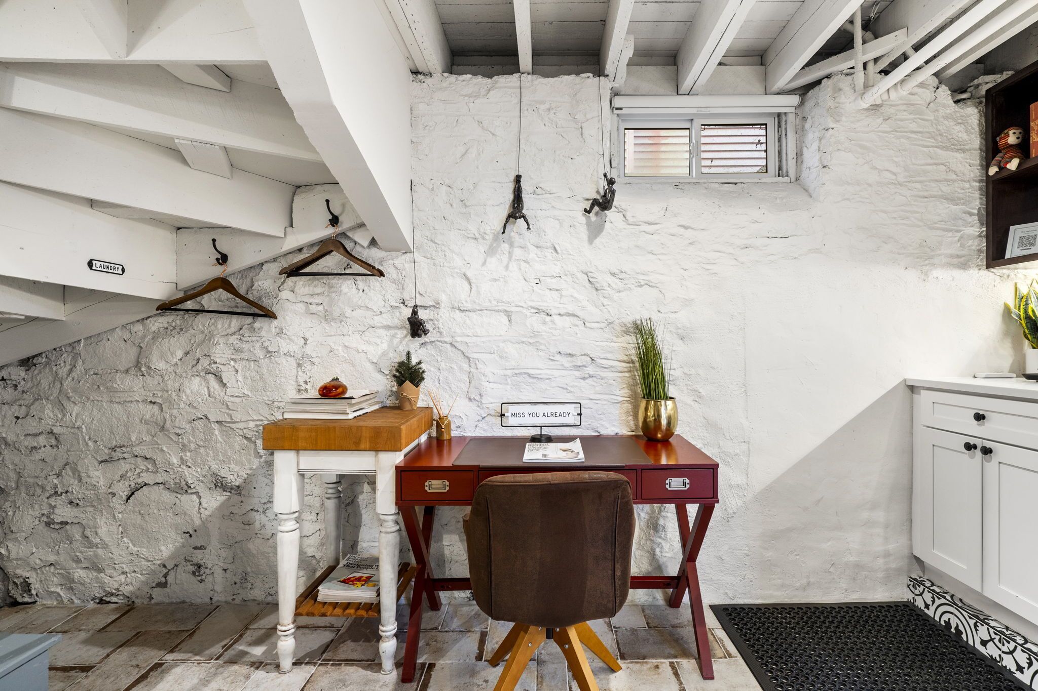Una habitación pequeña y rústica con una pared de ladrillo blanco, un escritorio de madera con una silla, una planta pequeña y un cartel que dice 'Miss You Already'.