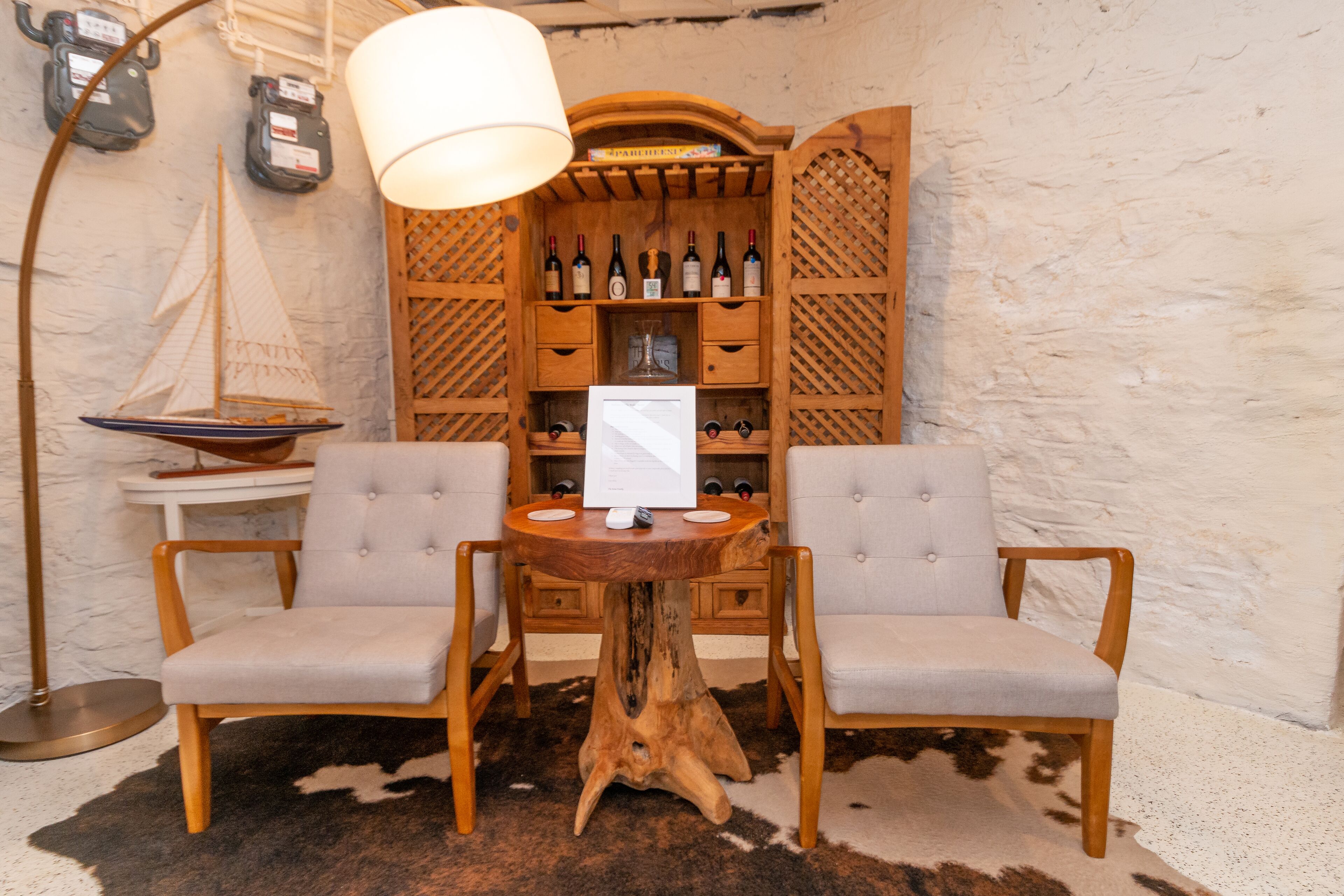 A wooden wine rack with bottles, two upholstered chairs, a table, and a ship model.