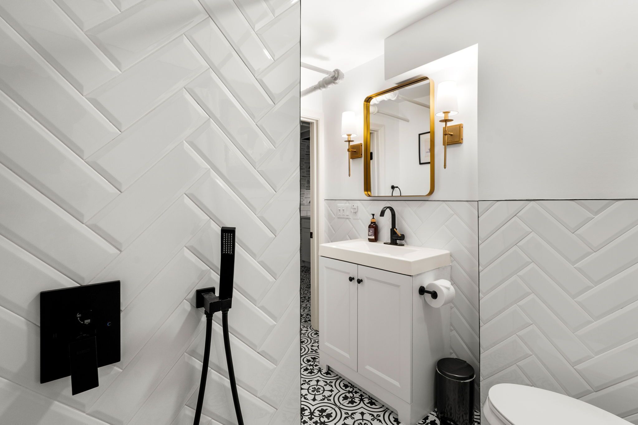 Un baño moderno con un patrón de baldosas en espiga, un lavamanos blanco con un espejo dorado y una estación de carga negra.