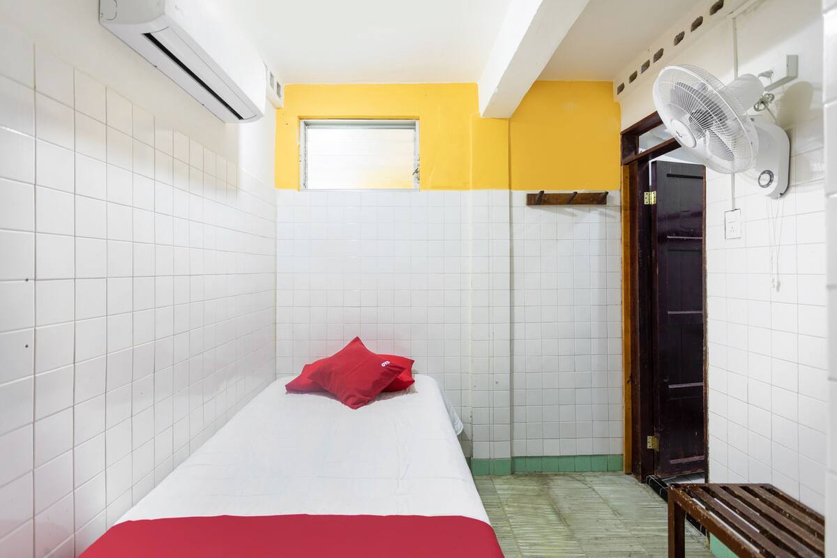A small, single-bed room with white and yellow walls, a red pillow, a wooden bench, and a wall-mounted fan.