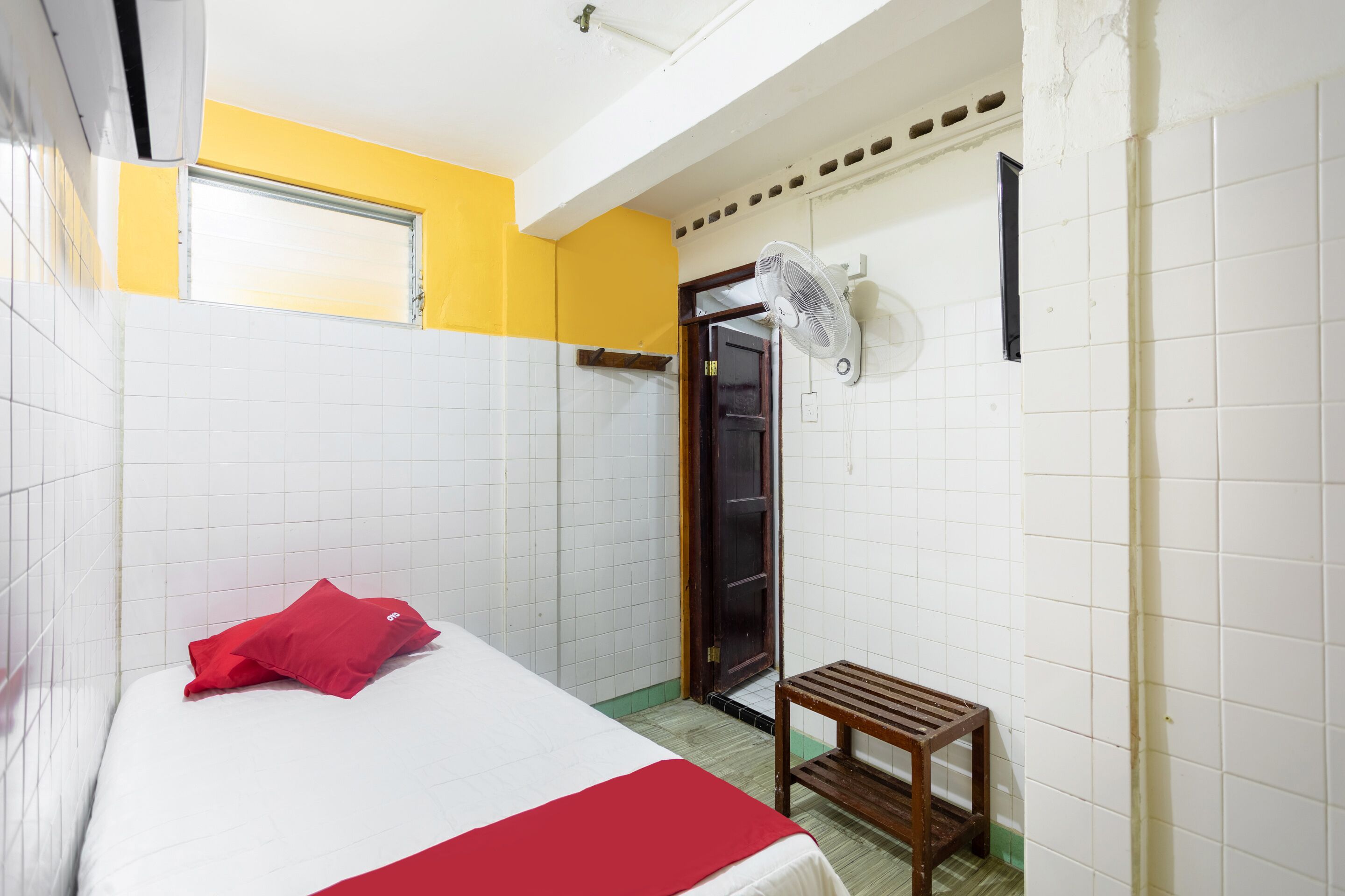 Habitación de hotel con cama, almohada roja, taburete de madera, ventilador y puerta que da al baño.