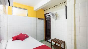 Habitación de hotel con cama, almohada roja, taburete de madera, ventilador y puerta que da al baño.
