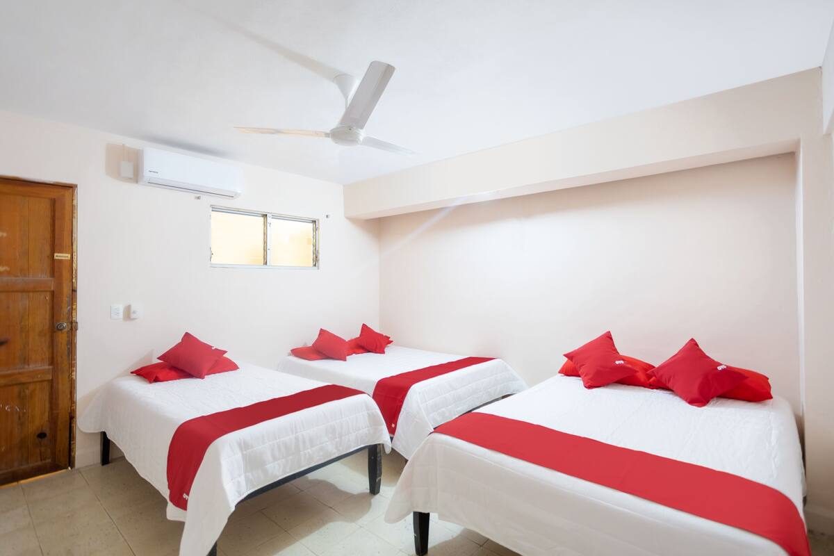 A room with two beds, each with red pillows and a white bedspread. There is a ceiling fan, an air conditioning unit, and a window.