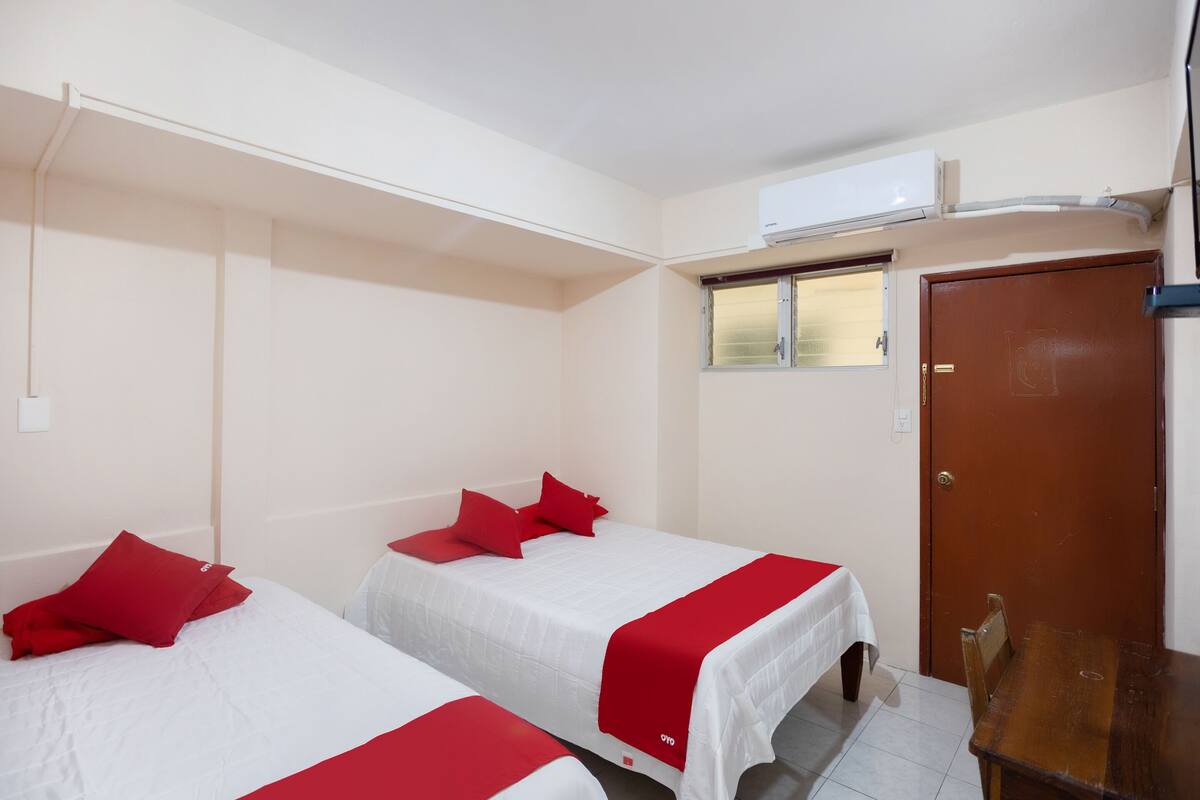 A hotel room with two beds, each with red bedding, a wooden table, and a window with blinds.
