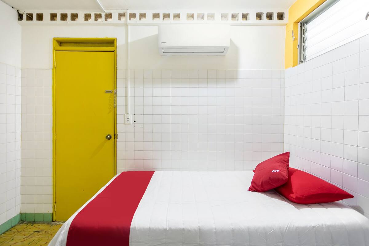 A small, brightly colored room with a yellow door, white tiled walls, and a bed with red pillows.