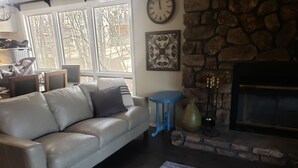 A living room with a stone fireplace, a leather sofa, a blue table, and a large window with a view of trees. - Tree house condo (Mt Ida)