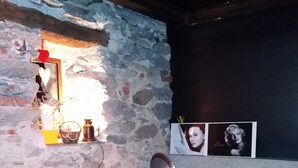 A room with a stone wall, a brown sofa with yellow cushions, a small table with a plant, and a wall-mounted shelf with decorative items. - Il Mondo In Valle (Pessinetto)