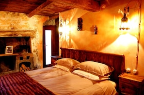 A bedroom with a wooden ceiling, a stone fireplace, a bed with white linens, and wall-mounted lights. - Il Mondo In Valle (Pessinetto)