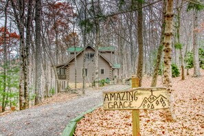 Eine rustikale Holzhütte mit Holzfassade und grünem Dach, umgeben von bewaldetem Gelände. Ein Kiesweg führt dorthin. Ein Schild davor zeigt 'AMAZING GRACE'.