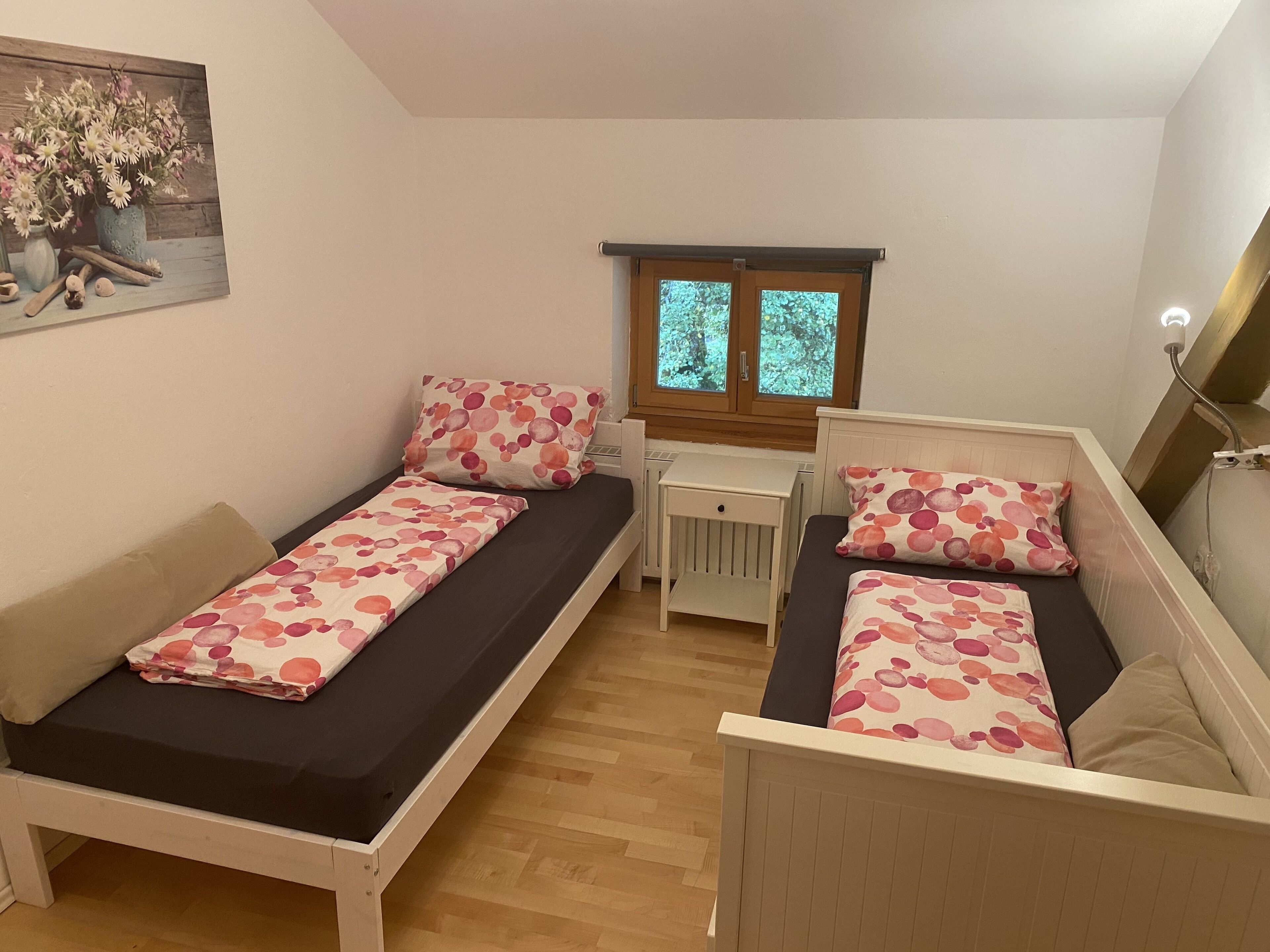 A small, cozy room with two single beds, a radiator, a window with a view of greenery, and a painting on the wall.