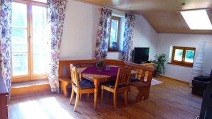 A cozy room with a wooden dining table, chairs, a TV, and a large window with curtains. - Vacation in the countryside with mountain views and close to the Chiemsee (Frasdorf)