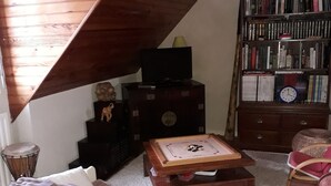 Ein gemütliches Dachzimmer mit schrägem Dach, einem hölzernen Bücherregal, einem kleinen Tisch mit einer Uhr und einer weißen Bank mit roter Decke.